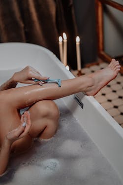 Woman in bathtub shaving her legs with a razor while sitting comfortably